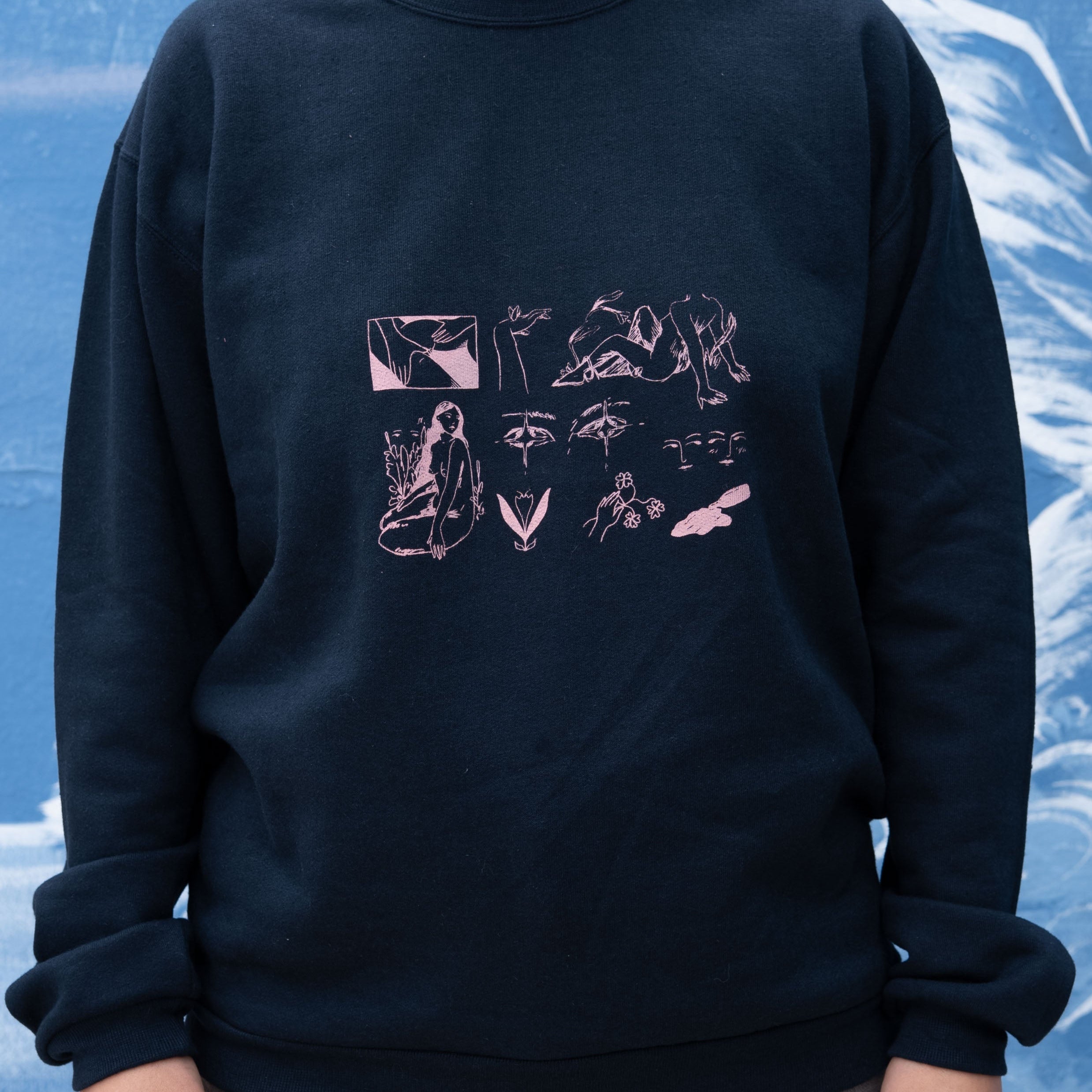 A person wearing a dark navy crewneck sweatshirt with a pink graphic print on the front, paired with gray jeans, set against a blue textured background.