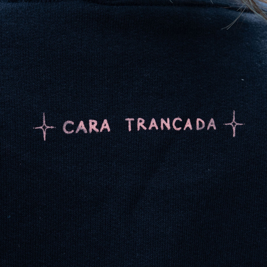 Close-up of a person wearing a black crewneck sweatshirt with 'CARA TRANCADA' embroidered in pink across the upper back.