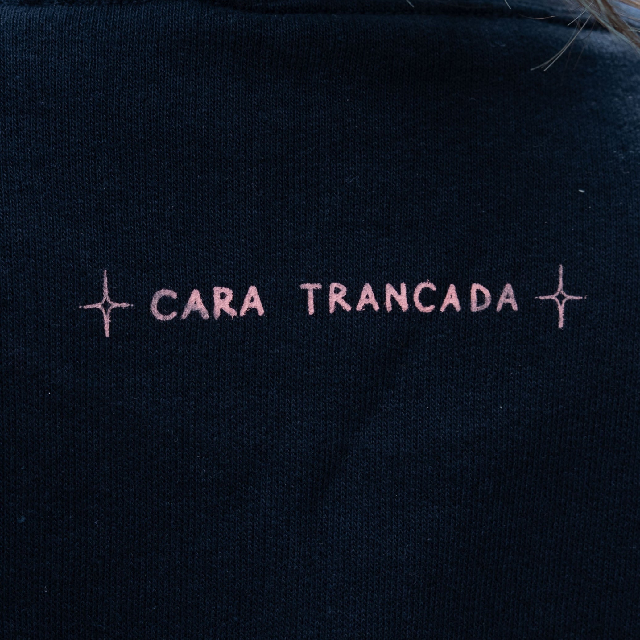 Close-up of a person wearing a black crewneck sweatshirt with 'CARA TRANCADA' embroidered in pink across the upper back.